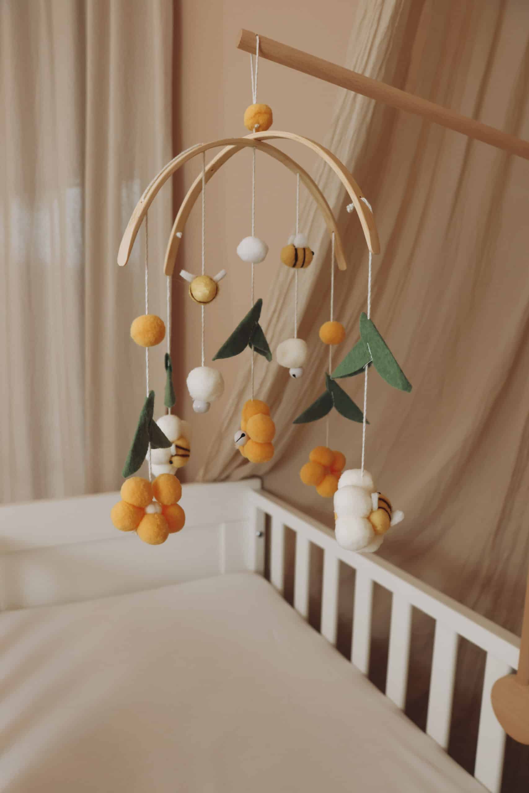 Mobile bébé en laine feutrée motifs fleurs jaunes, abeilles, feuilles vertes, suspendu au-dessus d’un lit blanc, décoration chambre enfant.